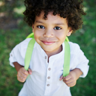 A toddler smiling into the camera with a white button-up and a green bookbag