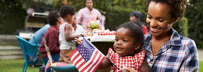 Mother and baby looks at American flag with rest of the family at picnic table behind them.