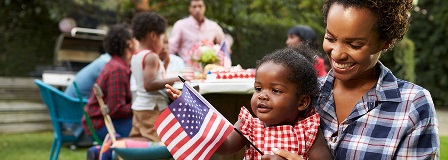 Mother and baby looks at American flag with rest of the family at picnic table behind them.