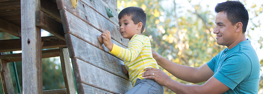 Father holds onto young son as he climbs wall at playground