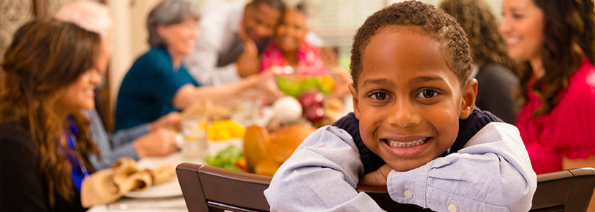 Little boy smiling at camera as family and friends gather around table to celebrate Thanksgiving